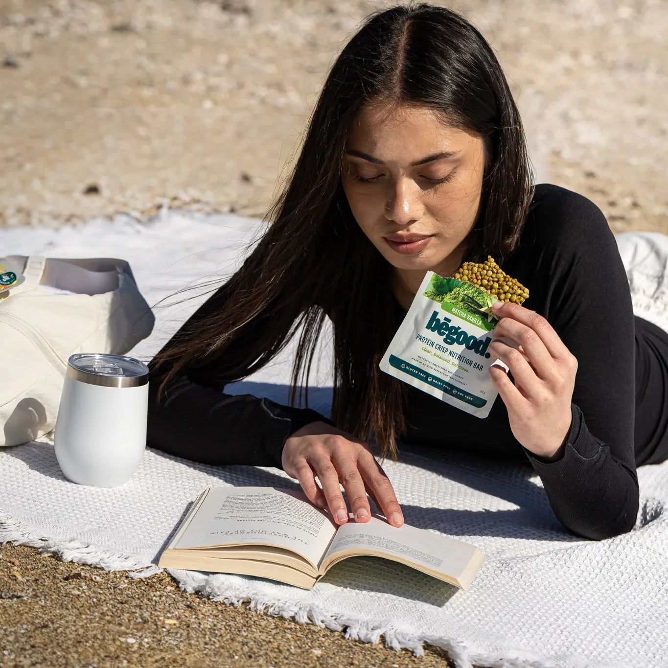 Woman lying on a blanket outdoors, reading a book and holding a bēgood nutrition product.