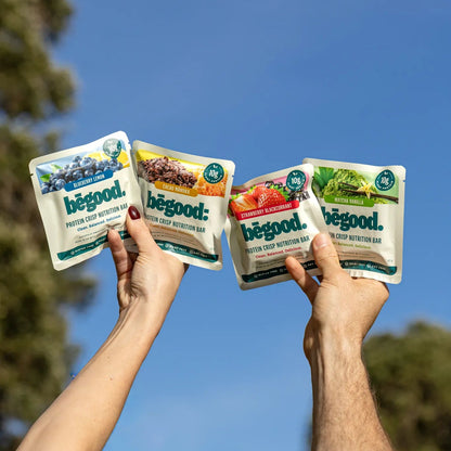 Two hands holding four packages of Begood protein bars against a blue sky with trees in the background