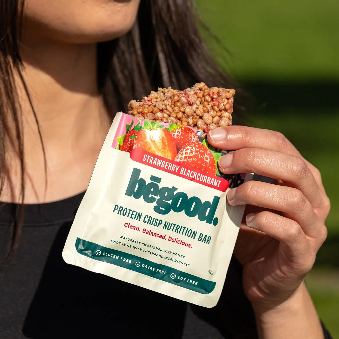Woman holding a 'begood' protein crisp nutrition bar with a green outdoor background