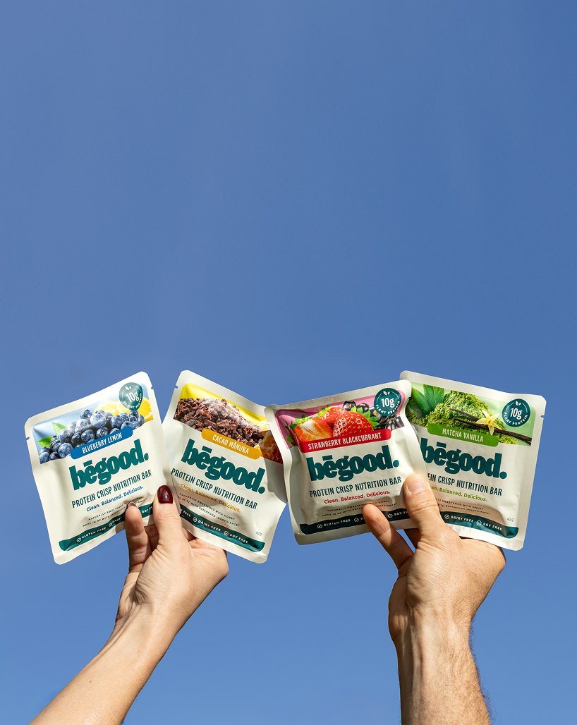 Four 'begood' protein crisp nutrition bar packages held against a blue sky.