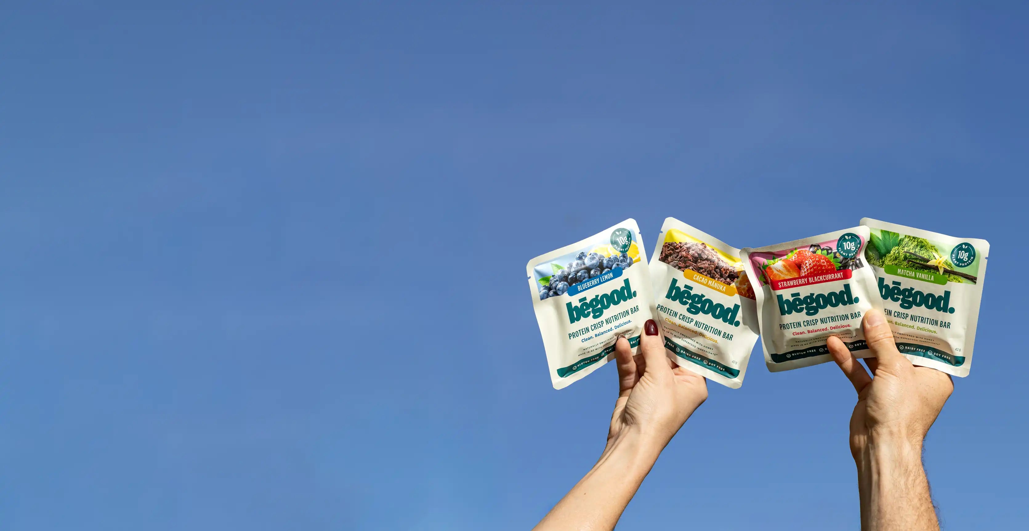 Two hands holding packages of 'begood' against a clear blue sky