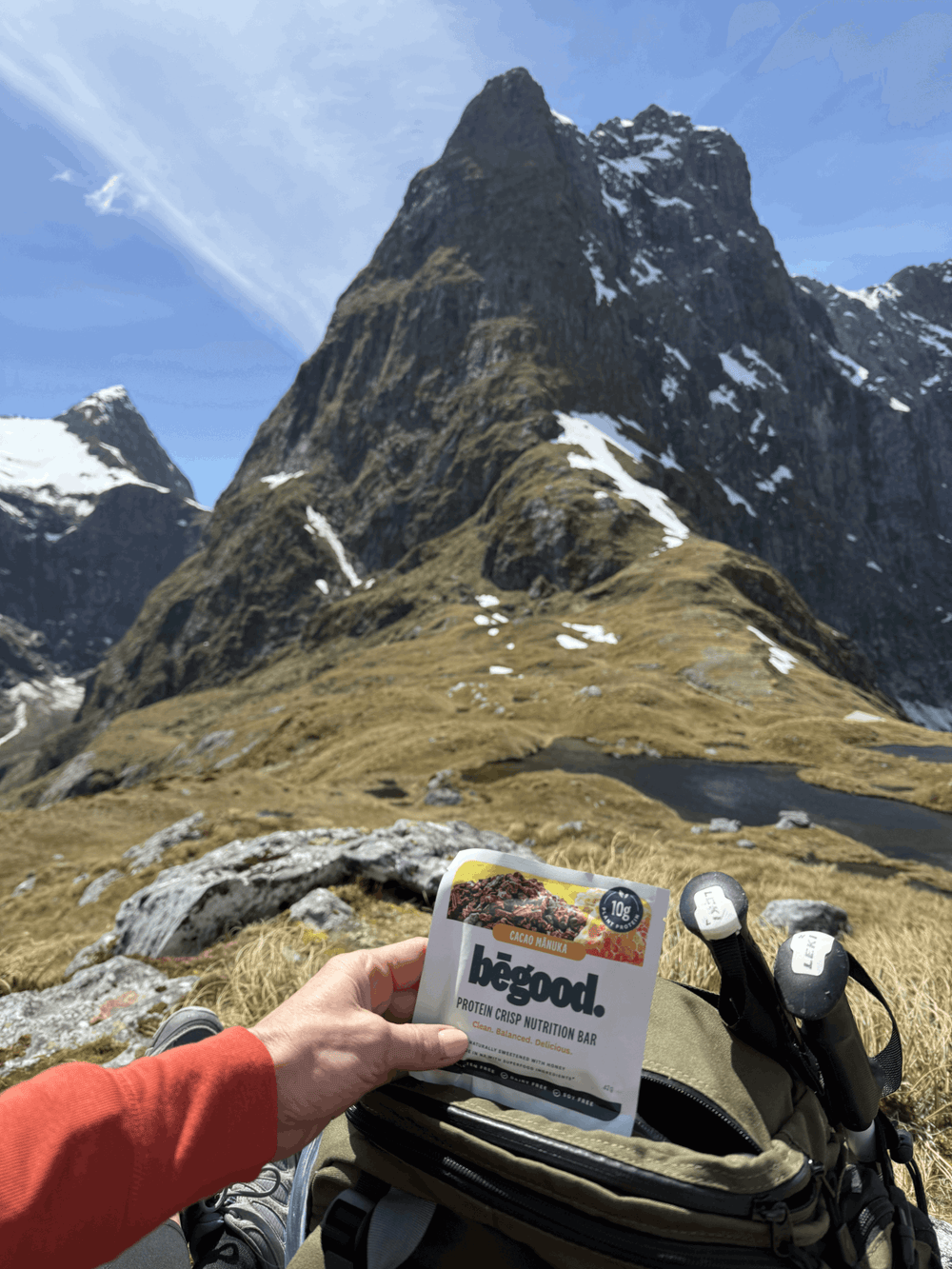 Person holding a 'hegood' protein bar with a mountainous landscape in the background on the Milford Great Walk in New Zealand