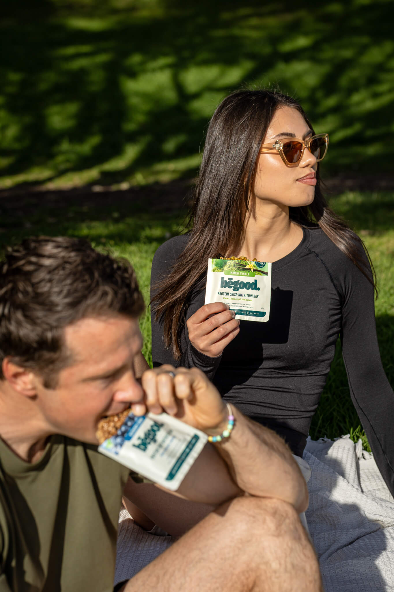Two people sitting outdoors, one eating Begood Nutrition Blueberry and the other holding a bar of Matcha Vanilla Bars