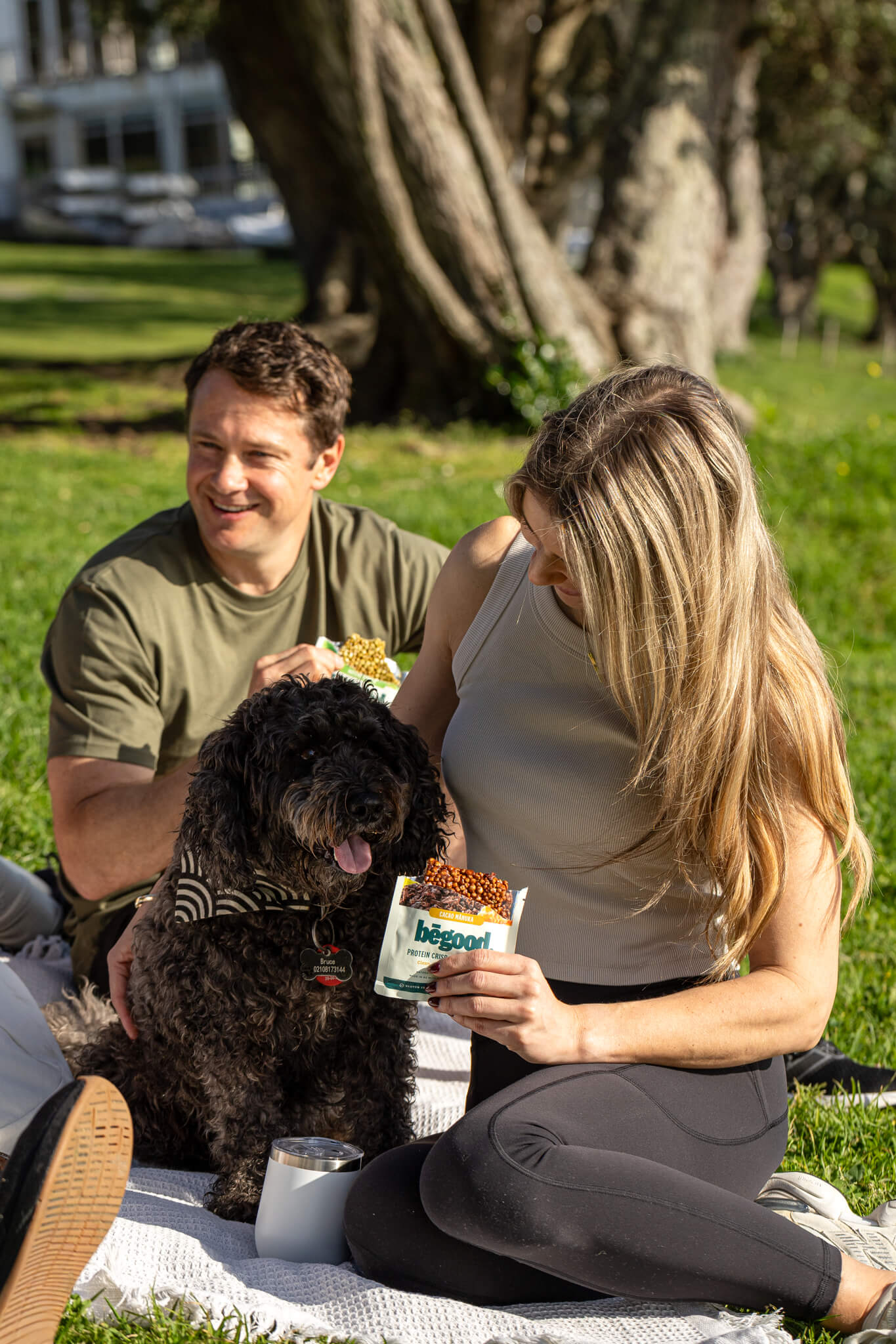 Man and woman sitting on a blanket with a dog in a park, eating Begood Nutrition Protein Crisp Bars.