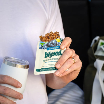 Person holding a package of 'begood' protein bars outdoors with a blurred background