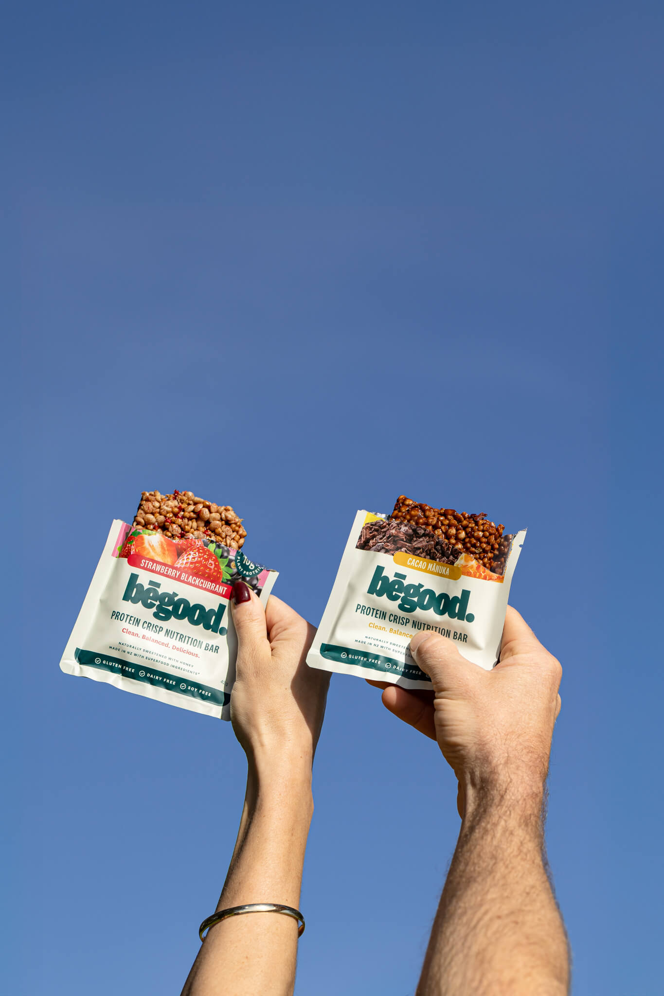 Two hands holding 'begood' protein crisp nutrition bars against a clear blue sky