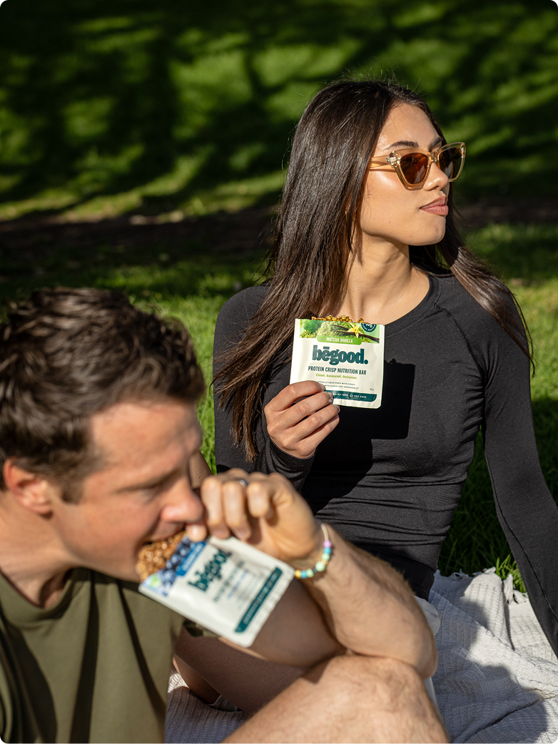 Two people sitting on grass, one eating a blueberry protein bar and the other holding a begood matcha protein bar.