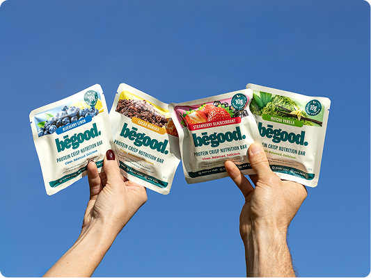 Four packages of 'begood' protein crisp nutrition bars held by two hands against a blue background